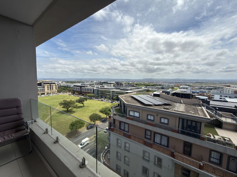 Elevated urban view from a balcony featuring a modern glass balustrade overlooking surrounding commercial buildings and green space.