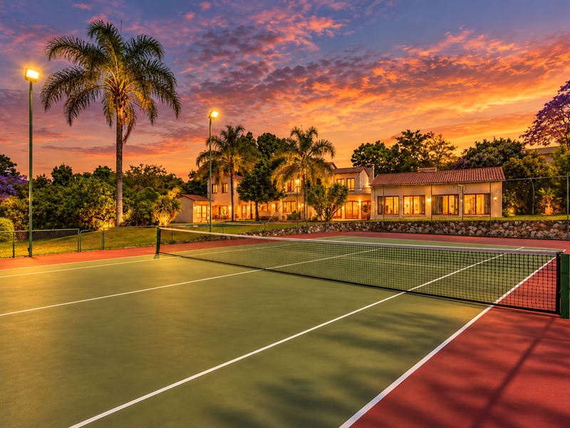A well-maintained private tennis court with floodlights, set against a beautifully landscaped garden and the main residence at sunset.