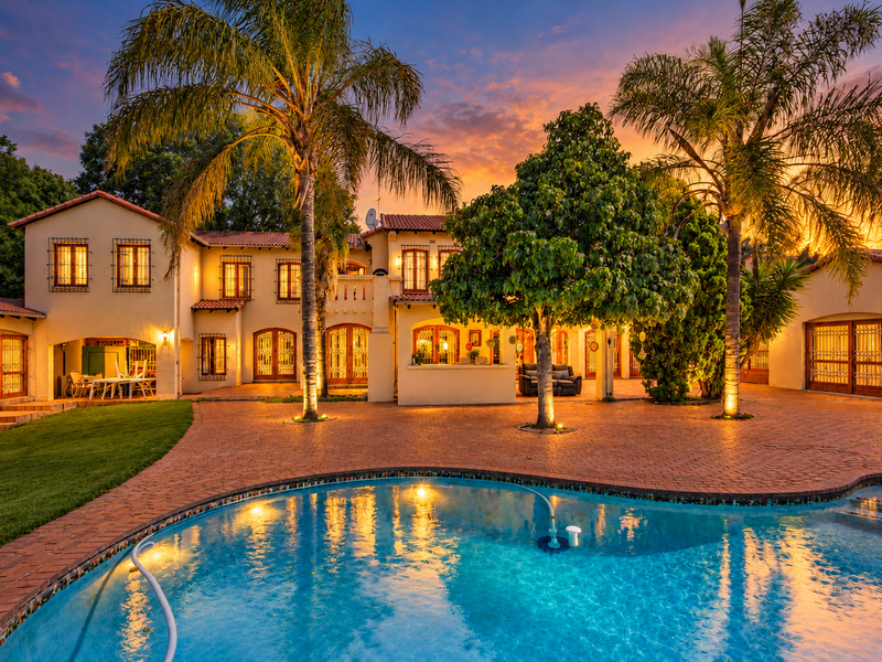 A stunning rear exterior view of a Mediterranean-style home featuring a large swimming pool, expansive brick-paved patio, and mature landscaping at sunset.