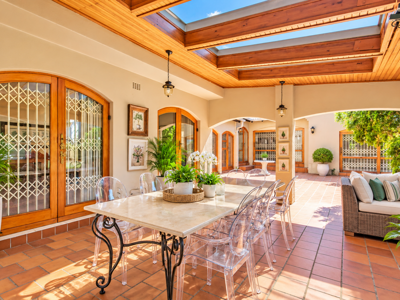 A spacious covered patio featuring terracotta flooring, a timber-paneled ceiling with a large skylight, and arched timber doors leading into the home.