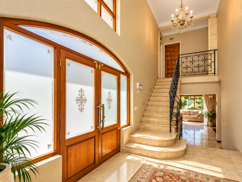 Grand entrance foyer featuring a double timber door with frosted glass, a tiled staircase with wrought iron balustrade, and an elegant chandelier.