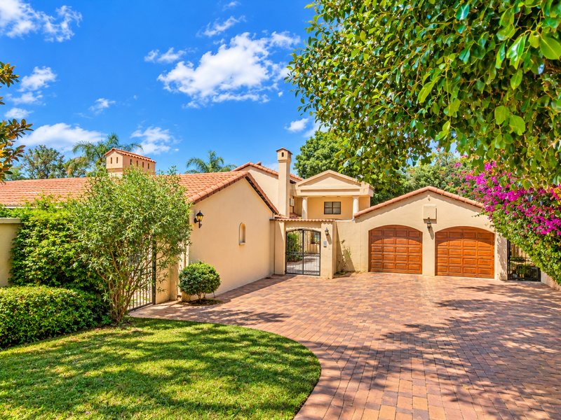 A well-maintained front exterior featuring a paved driveway, double garage, and established garden with terracotta roof tiles and a secure gated entrance.