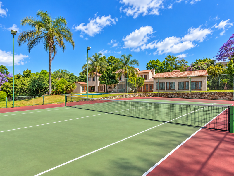 A well-maintained private tennis court featuring a hard surface, perimeter fencing, and integrated floodlights, set within a landscaped residential garden.