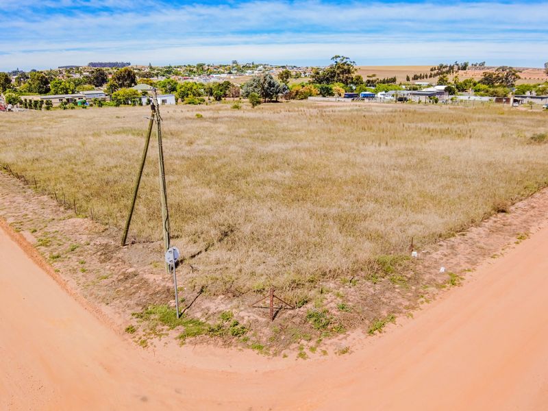 An elevated aerial view of a vacant residential plot situated on a corner lot with dirt road access and existing utility infrastructure.