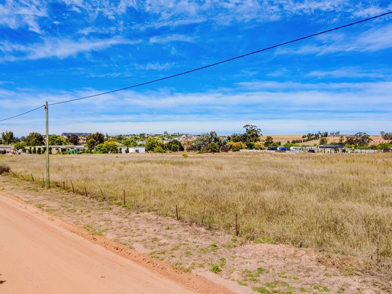 A vacant residential plot of land featuring a dirt road frontage, wire fencing, and utility infrastructure, offering an open landscape with surrounding neighborhood views.