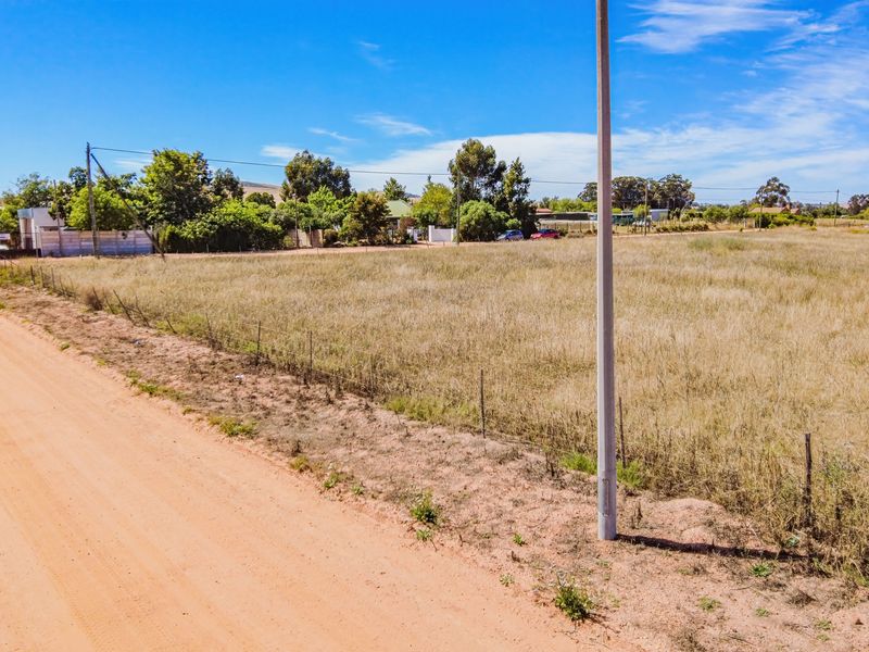 A vacant residential plot of land featuring dry grass, bordered by a dirt road and wire fencing with a utility pole on site.