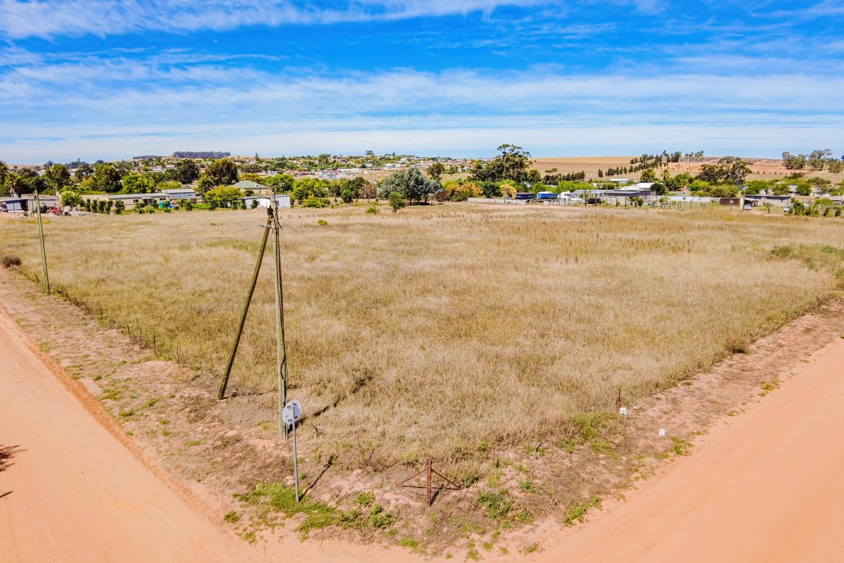 An aerial view of a vacant residential plot situated at a corner, featuring dry grass, perimeter fencing, and access to dirt roads.