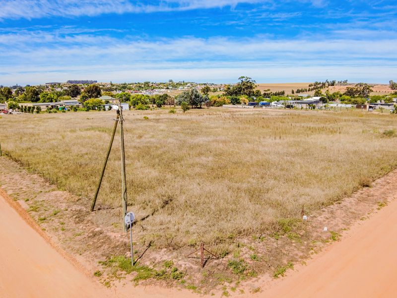 An aerial view of a vacant residential plot situated at a corner, featuring dry grass, perimeter fencing, and access to dirt roads.