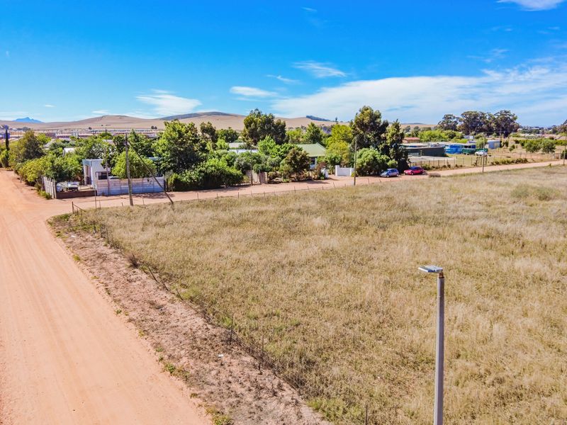 Aerial view of a vacant residential plot adjacent to a dirt road, featuring perimeter wire fencing and a street light.