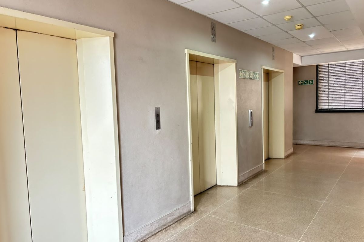 A functional apartment building hallway featuring multiple elevator access points, tiled flooring, and a suspended ceiling with integrated lighting.