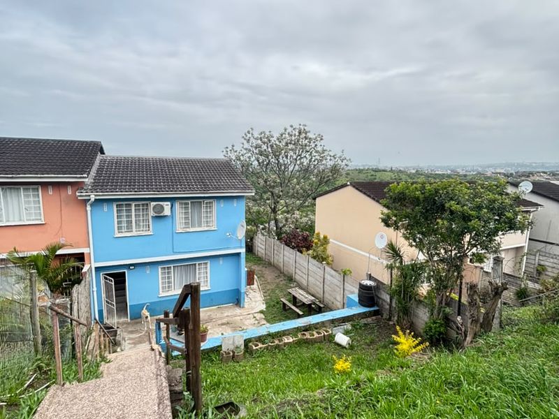 Rear view of a blue two-story house featuring a concrete staircase, patio area, and precast boundary wall, set within a sloped garden.