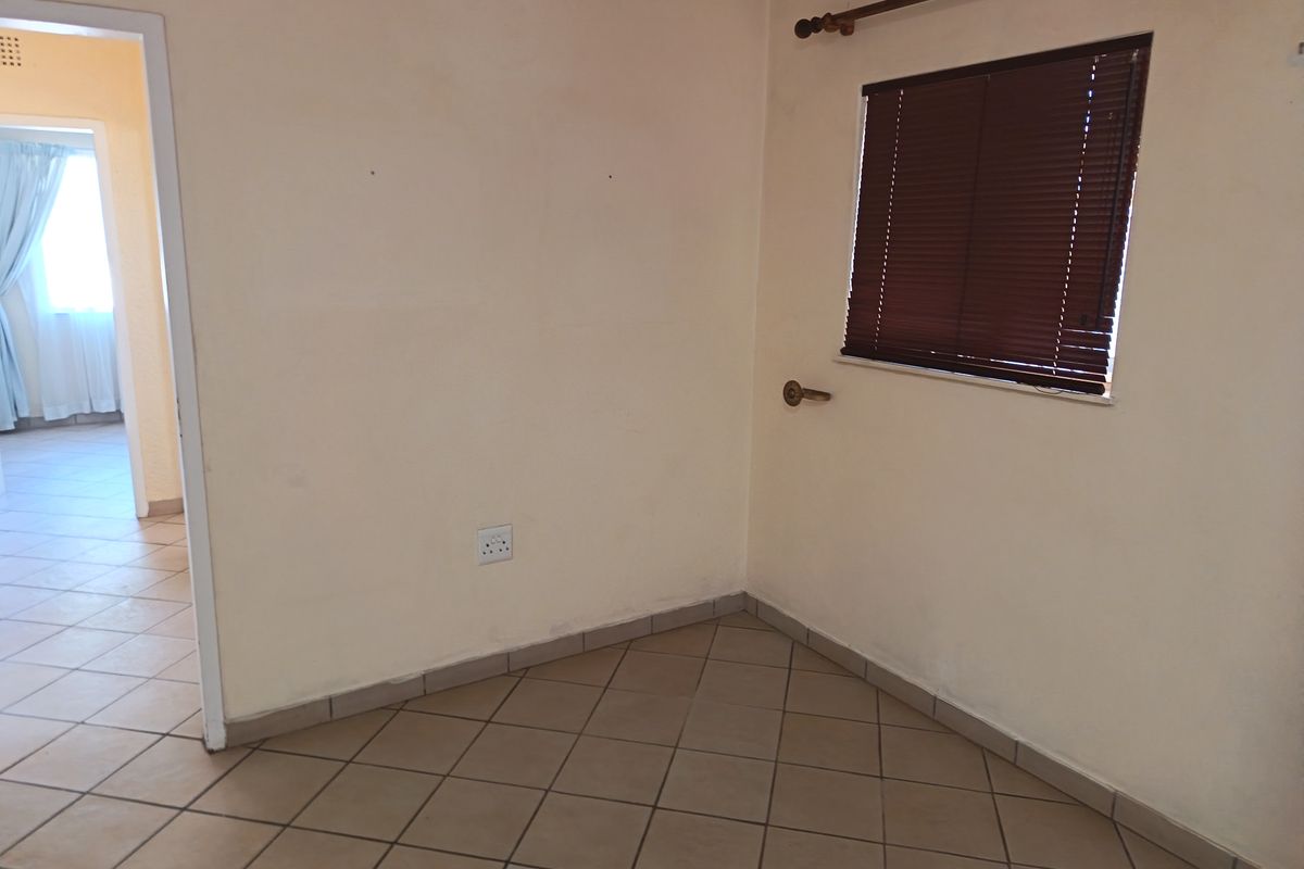 An empty bedroom featuring neutral tiled flooring, a window with fitted venetian blinds, and a doorway leading to an adjacent room.