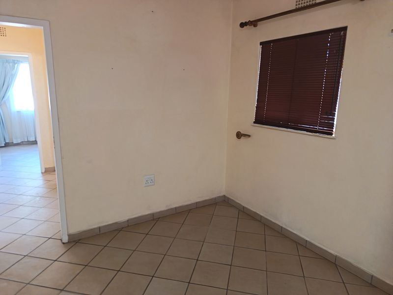 An empty bedroom featuring neutral tiled flooring, a window with fitted venetian blinds, and a doorway leading to an adjacent room.