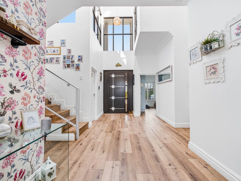 Spacious double-volume entrance hall featuring timber flooring, a modern staircase with glass balustrade, and a striking feature wallpaper wall.