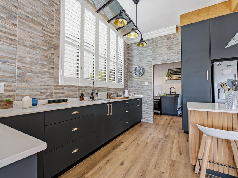 Modern kitchen featuring dark cabinetry, stone-look wall tiles, quartz countertops, plantation shutters, and an adjoining scullery area with wood-look flooring.