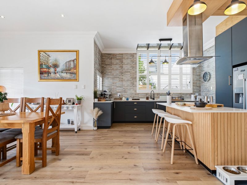A modern open-plan kitchen and dining area featuring a timber-clad island, stone countertops, dark cabinetry, and high-quality timber flooring with plantation shutters.