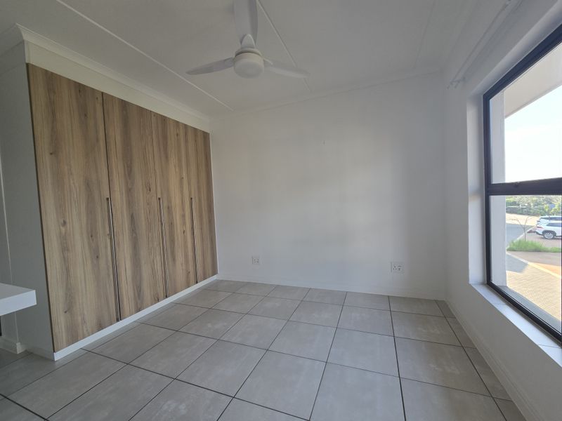 A bright, empty bedroom featuring modern wood-finish built-in cupboards, neutral tiled flooring, and a ceiling fan, offering a clean and versatile living space.