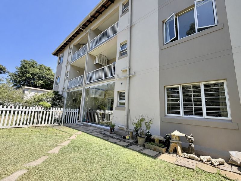 Rear view of a multi-story apartment building featuring private balconies, sliding glass doors, and a well-maintained garden area with a white picket fence.