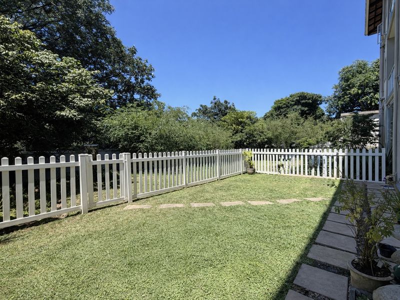 A well-maintained rear garden featuring a white picket fence, manicured lawn, and stone pathway leading along the side of the apartment building.