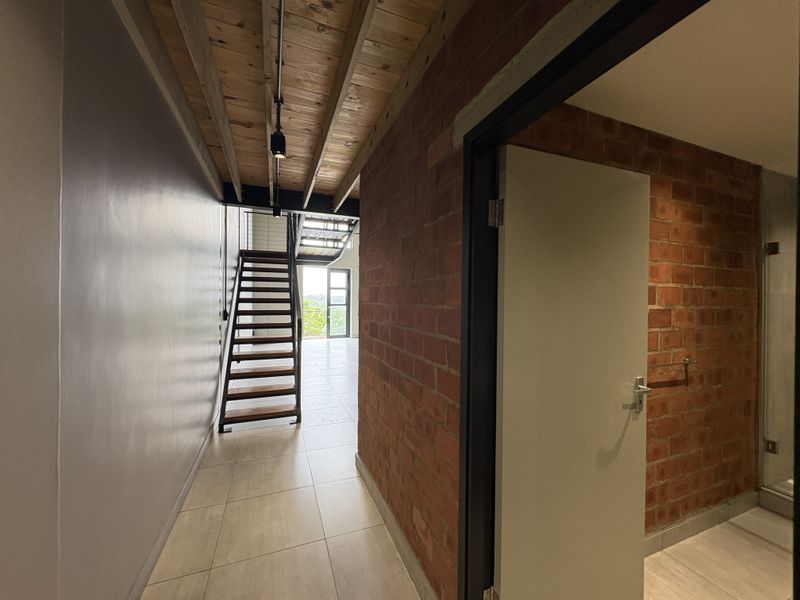 A modern hallway featuring exposed timber ceiling beams, industrial-style brickwork, and a staircase leading to an upper level with tiled flooring throughout.