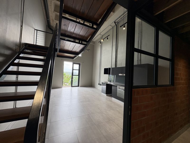 Modern open-plan living area featuring industrial design elements, a sleek kitchen, exposed brickwork, and a striking staircase leading to an upper level.