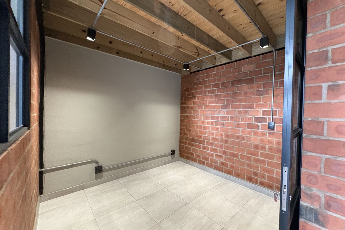An empty industrial-style room featuring exposed brick walls, timber ceiling beams, track lighting, and modern tiled flooring.