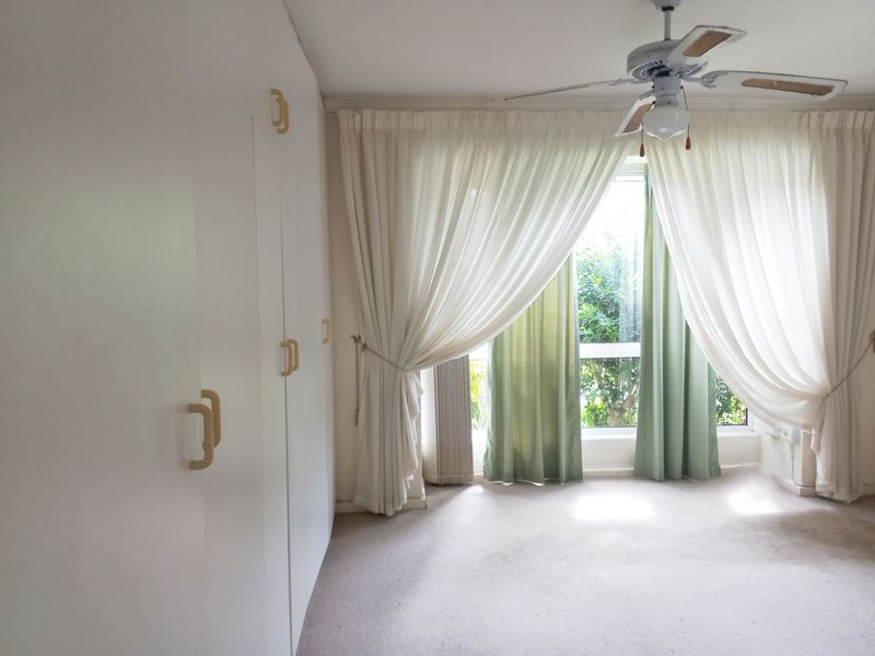 A spacious, empty bedroom featuring built-in cupboards, a ceiling fan, and large windows with curtains, offering a bright and neutral canvas for renovation.