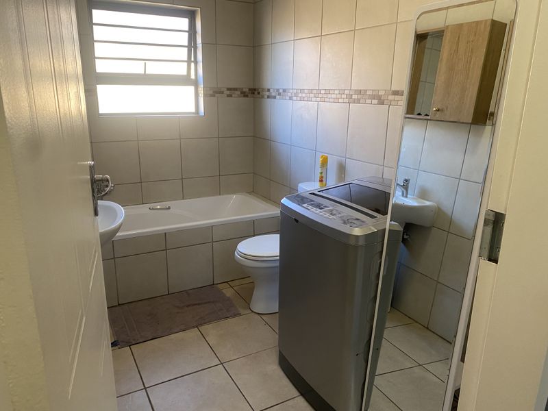 A functional bathroom featuring a bathtub, toilet, and wall-mounted basin with neutral tiled finishes and a louvered window for ventilation.