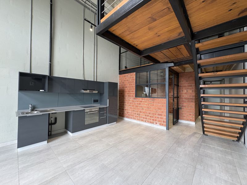 Modern industrial-style open plan kitchen and living area featuring high ceilings, exposed brickwork, a mezzanine level, and sleek dark cabinetry.