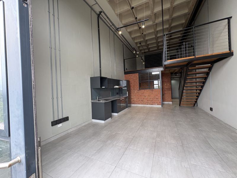 Modern industrial-style open plan apartment featuring a double-volume space, exposed concrete waffle ceiling, mezzanine level, and a sleek kitchen with exposed brick accents.