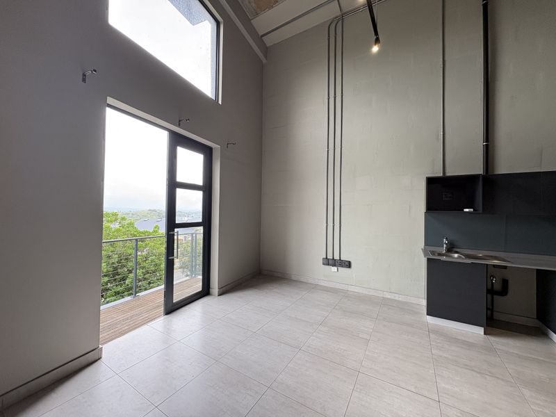 Modern open-plan studio apartment featuring high ceilings, exposed industrial-style conduit, tiled flooring, a compact kitchenette, and direct access to a private balcony.