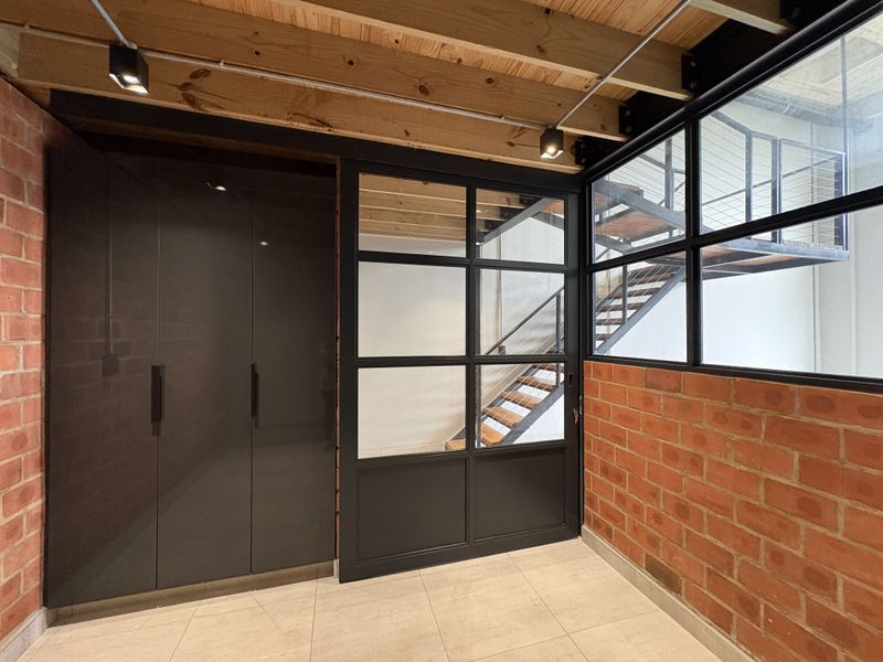 Modern industrial-style hallway featuring exposed brick walls, timber ceiling beams, sleek built-in cabinetry, and a striking steel-framed glass partition overlooking a staircase.