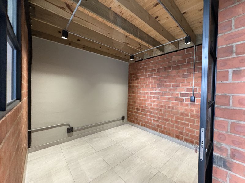 An empty industrial-style room featuring exposed brick walls, timber ceiling beams, track lighting, and modern tiled flooring.