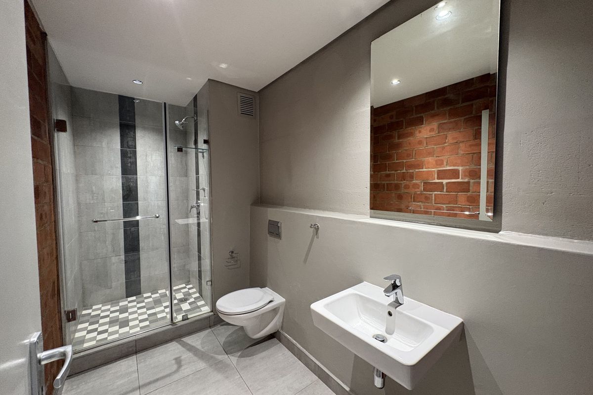 Modern bathroom featuring a glass-enclosed shower, wall-mounted toilet, basin, and an industrial-style exposed brick feature wall with contemporary grey tiling.