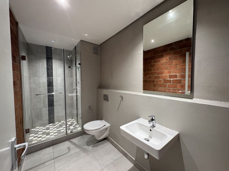 Modern bathroom featuring a glass-enclosed shower, wall-mounted toilet, basin, and an industrial-style exposed brick feature wall with contemporary grey tiling.