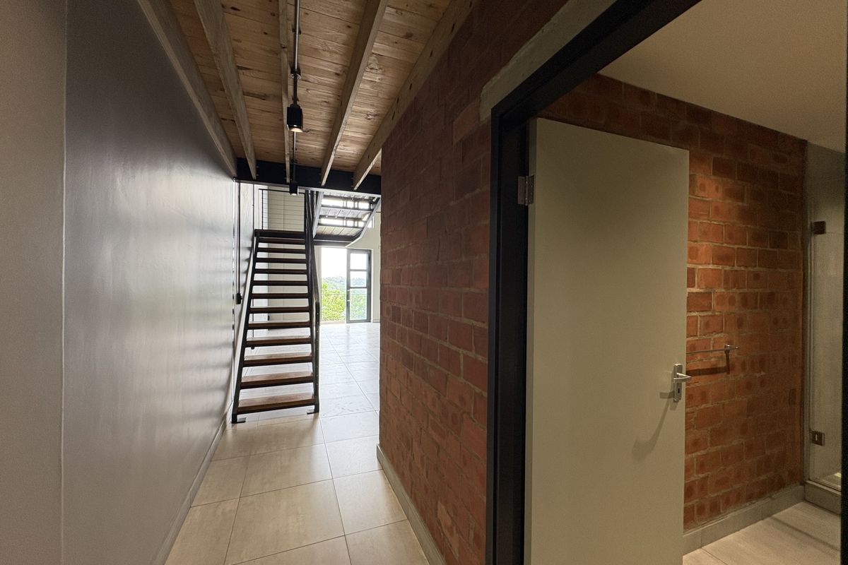 A modern hallway featuring exposed timber ceiling beams, industrial-style brickwork, and a staircase leading to an upper level with tiled flooring throughout.