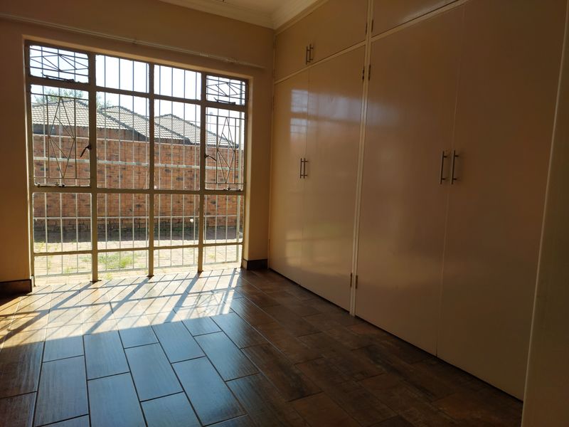 A bright, empty bedroom featuring modern wood-look tiled flooring, extensive built-in storage cupboards, and large windows with security bars.