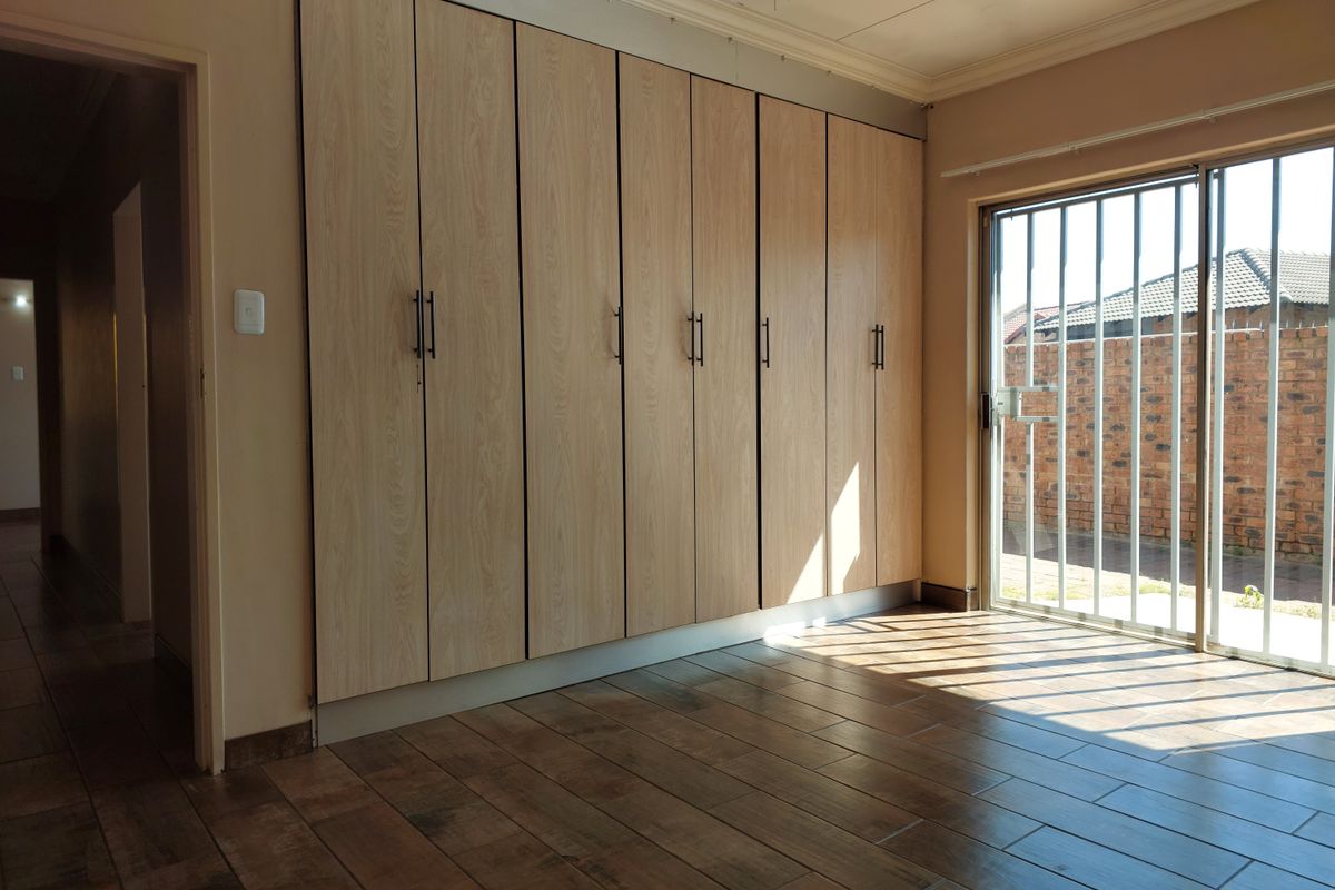 A bright, empty bedroom featuring modern wood-look tiled flooring, extensive built-in cupboards, and a sliding door with security gate leading to the exterior.