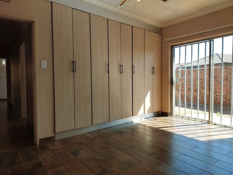 A bright, empty bedroom featuring modern wood-look tiled flooring, extensive built-in cupboards, and a sliding door with security gate leading to the exterior.