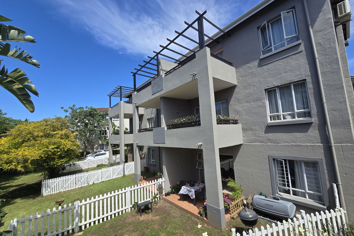 Rear view of a multi-story apartment building featuring private balconies with pergolas, ground-floor patio access, and a fenced garden area.