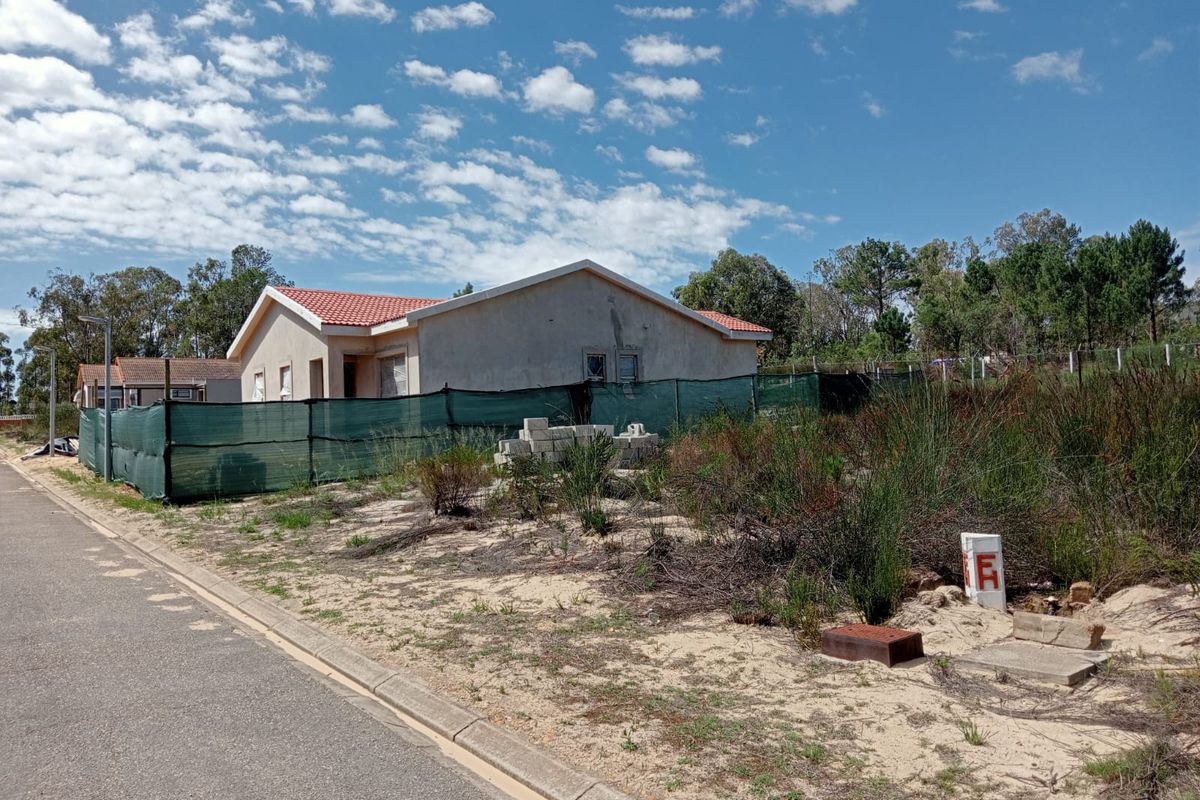 A view of a residential vacant land plot featuring a nearby house, sandy terrain, and utility infrastructure along the street.