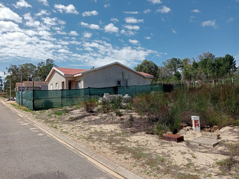 A view of a residential vacant land plot featuring a nearby house, sandy terrain, and utility infrastructure along the street.