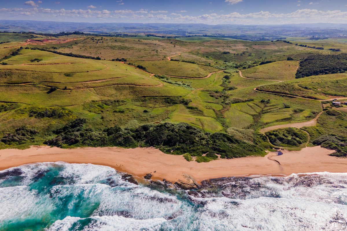 An expansive aerial view of a coastal residential land parcel featuring rolling green hills, natural vegetation, and direct access to a sandy beach.