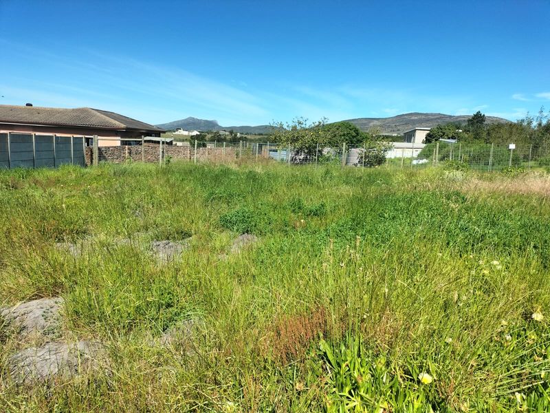 A vacant residential plot featuring overgrown grass, enclosed by wire mesh fencing and a stone boundary wall with mountain views in the background.