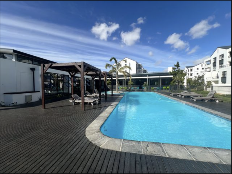 A well-maintained communal swimming pool area featuring timber decking, a shaded pergola structure, and stone coping, set within an apartment complex.