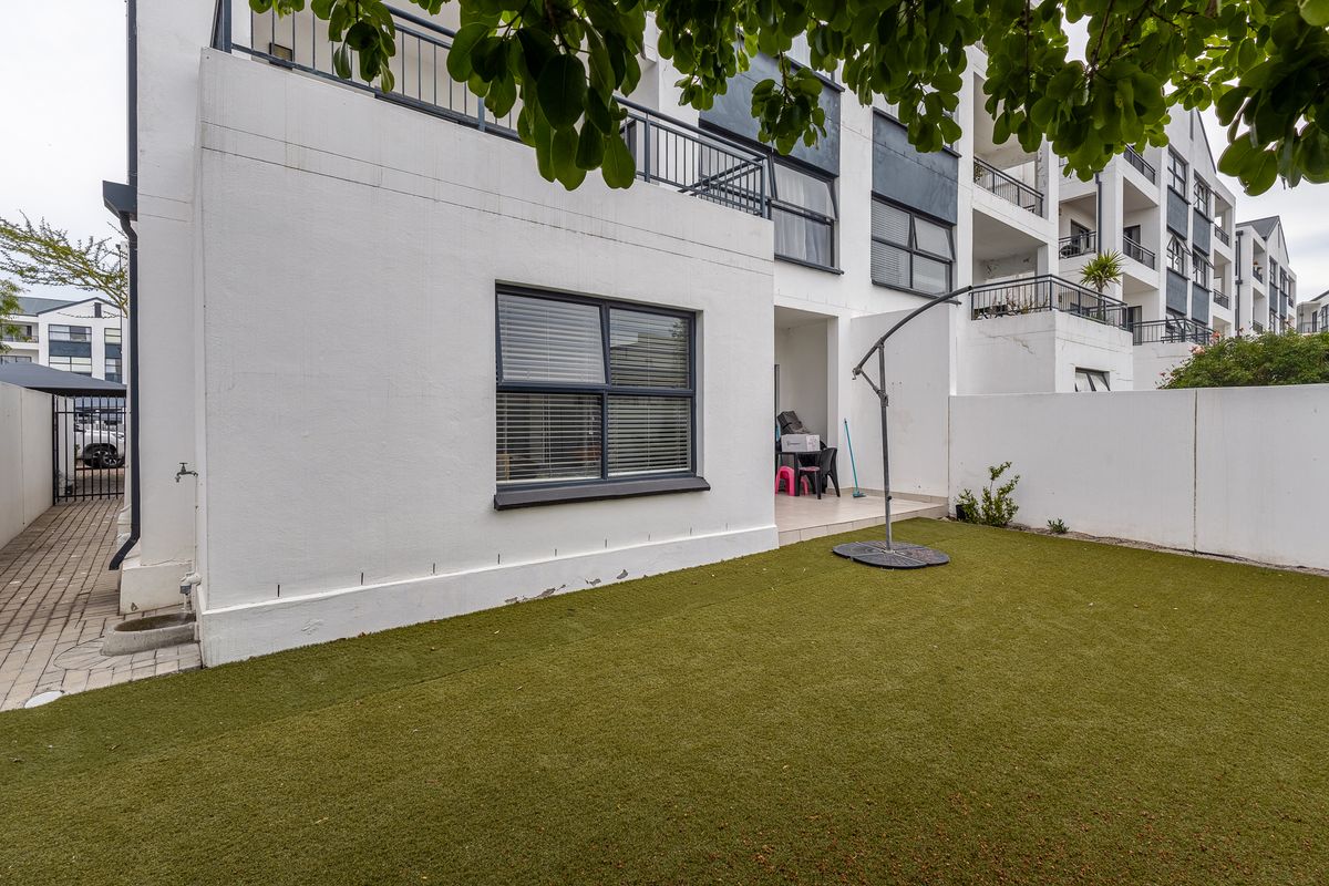Rear view of the apartment featuring a private garden with artificial lawn, a paved walkway, and a balcony with metal railings.