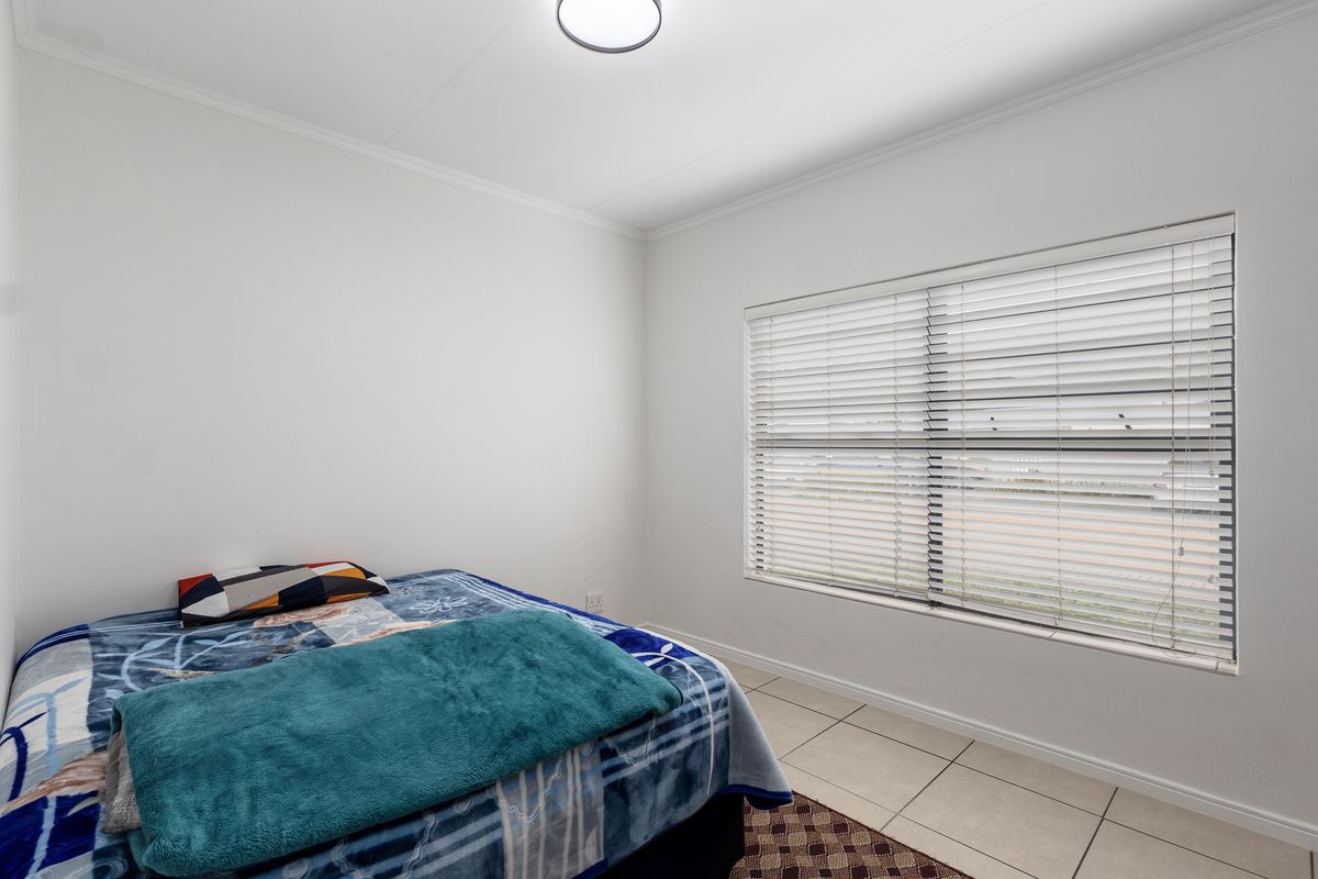 A clean and bright bedroom featuring neutral tiled flooring, a large window with fitted venetian blinds, and simple ceiling lighting.