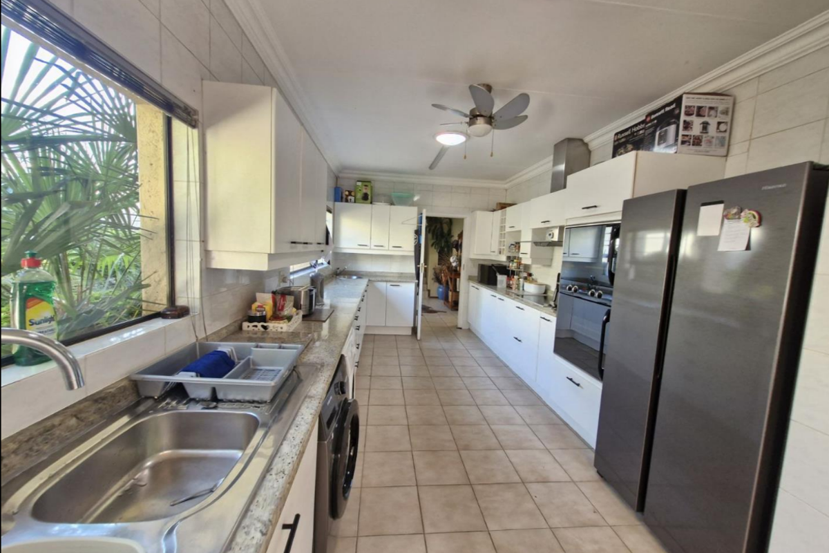 A functional galley-style kitchen featuring white cabinetry, stone-look countertops, stainless steel double sink, built-in oven, and tiled flooring throughout.