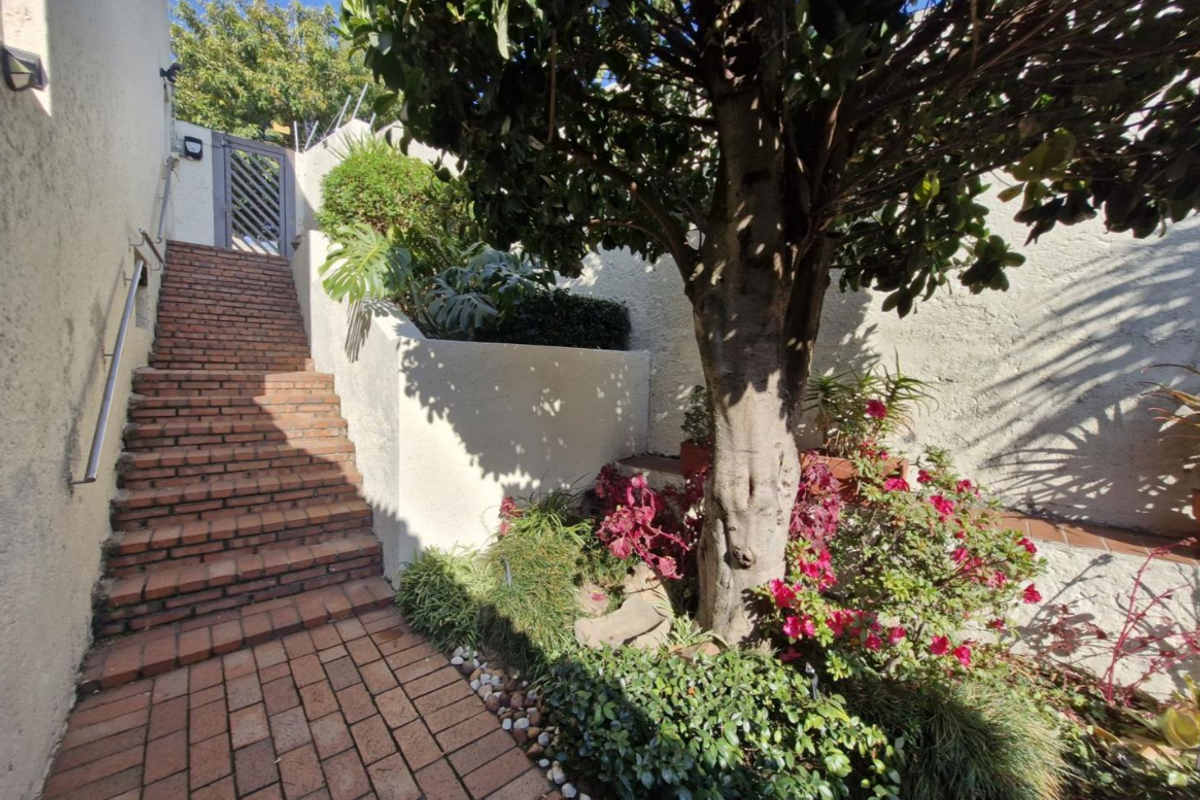 An outdoor staircase featuring brick steps and a metal handrail, leading up to a gated entrance surrounded by established garden landscaping.