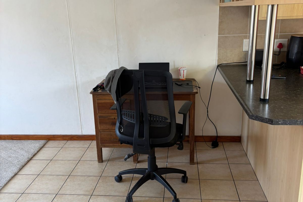 A functional kitchen area featuring a dark laminate countertop, light wood cabinetry, and tiled flooring, currently utilized as a workspace.